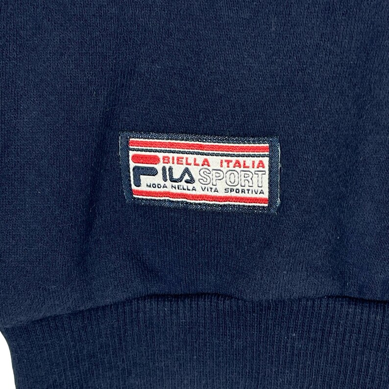 May include: A dark navy blue fabric garment featuring a rectangular patch with red and white horizontal stripes. The patch displays the text BIELLA ITALIA, FILA SPORT, and MODA NELLA VITA SPORTIVA in black and red lettering on a white background.