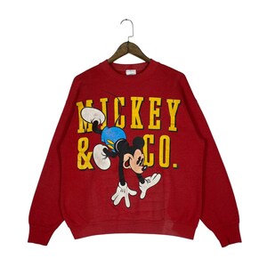 May include: Red sweatshirt featuring Mickey Mouse in a playful pose. The graphic includes the text "MICKEY & CO." in yellow block letters. Mickey is depicted in his classic attire: white gloves, blue shorts, and black ears.