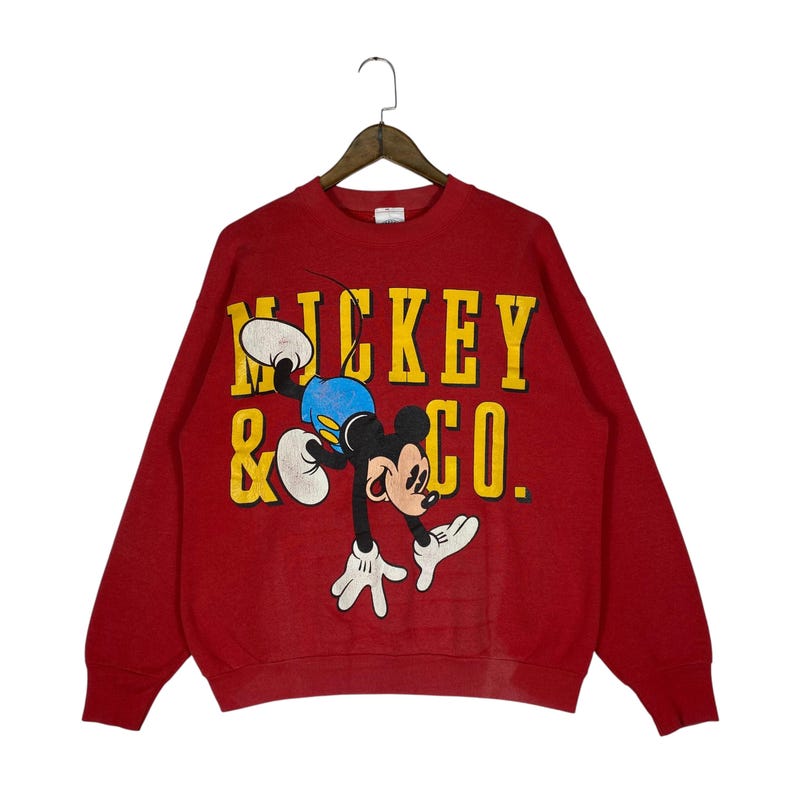 May include: Red sweatshirt featuring Mickey Mouse in a playful pose. The graphic includes the text "MICKEY & CO." in yellow block letters. Mickey is depicted in his classic attire: white gloves, blue shorts, and black ears.