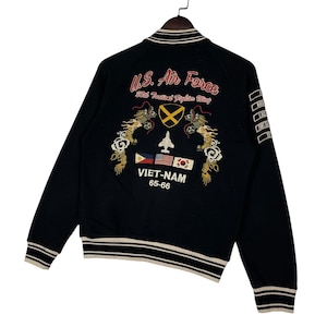 May include: Black bomber jacket with embroidered details. Features "U.S. Air Force" text, a jet, flags, and dragon designs. The jacket has white and beige striped trim on the collar, cuffs, and hem. The text "VIET-NAM 65-66" is also visible.