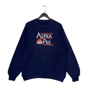 May include: A navy blue crewneck sweatshirt featuring the white embroidered text Alpha Phi, a red circular graphic, and a tan cowboy hat icon. The design includes two horizontal red bars above and below the text, with small white text below.
