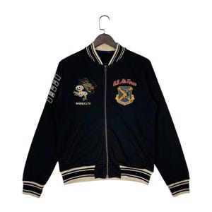May include: Black bomber jacket with embroidered dragon and U.S. Air Force patches. The jacket has a zip-up front, striped cuffs, and the text "AVIREX LTD". A classic, vintage-inspired outerwear piece.