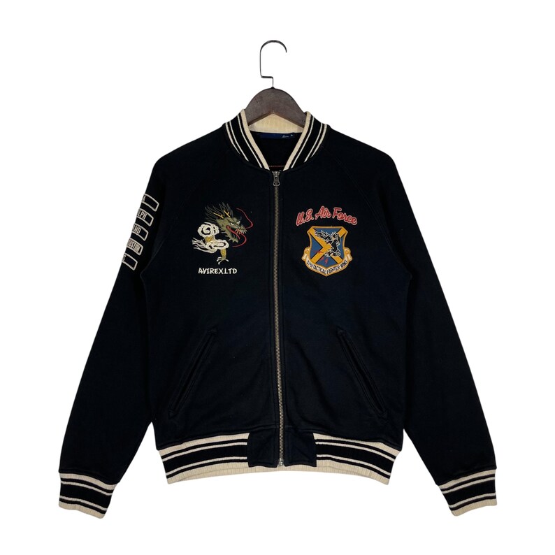 May include: Black bomber jacket with embroidered dragon and U.S. Air Force patches. The jacket has a zip-up front, striped cuffs, and the text "AVIREX LTD". A classic, vintage-inspired outerwear piece.