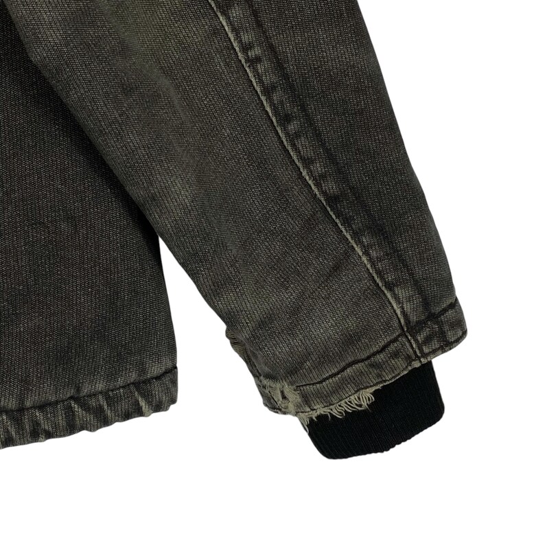 May include: Close-up of a dark grey denim jacket sleeve. The sleeve has a ribbed black cuff. The denim shows signs of wear and fading, with visible stitching and a slightly distressed appearance. The jacket appears to be vintage or pre-worn.