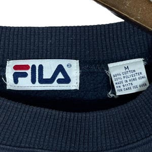 Puede incluir: Primer plano de una sudadera FILA azul marino. La etiqueta blanca muestra el logotipo de la marca en rojo y azul. Una etiqueta m&aacute;s peque&ntilde;a indica que la sudadera es talla M, hecha de algod&oacute;n y poli&eacute;ster, y fabricada en Hong Kong.