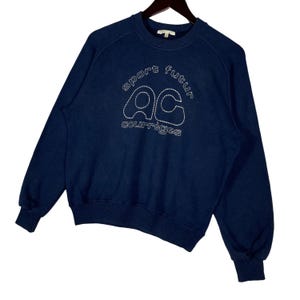 May include: A navy blue sweatshirt featuring white embroidered text. The text reads "sport futur" above the letters "ac" and "courr&egrave;ges" below. The sweatshirt has a crew neck and long sleeves with ribbed cuffs.