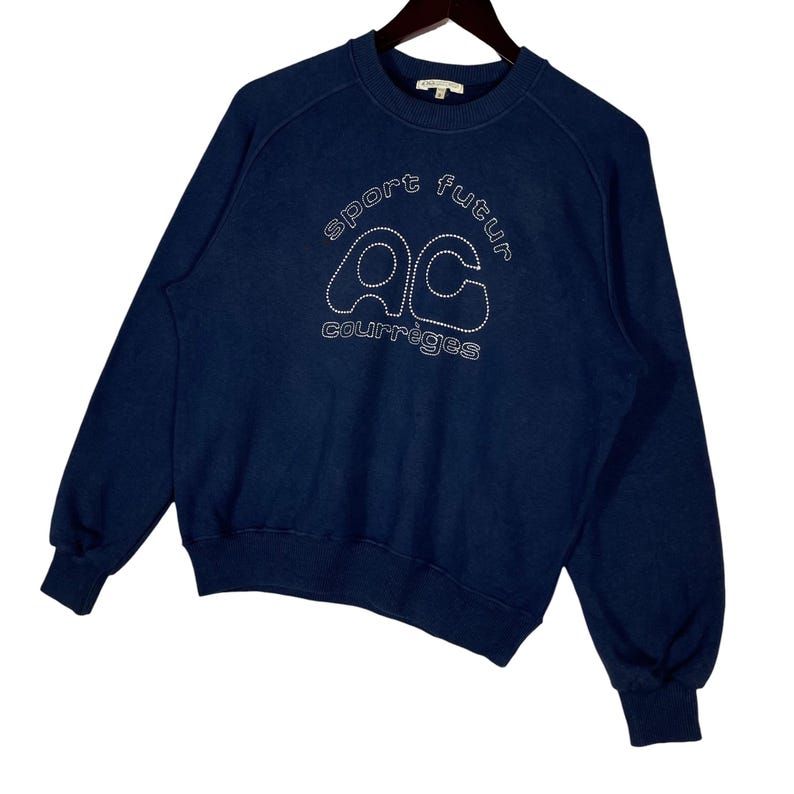 May include: A navy blue sweatshirt featuring white embroidered text. The text reads "sport futur" above the letters "ac" and "courr&egrave;ges" below. The sweatshirt has a crew neck and long sleeves with ribbed cuffs.