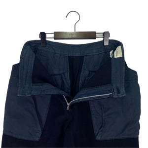 May include: Dark navy blue pants with a contrasting fabric panel on the front. The pants are hanging on a wooden hanger. The zipper is open, and a small tag is visible on the right side.