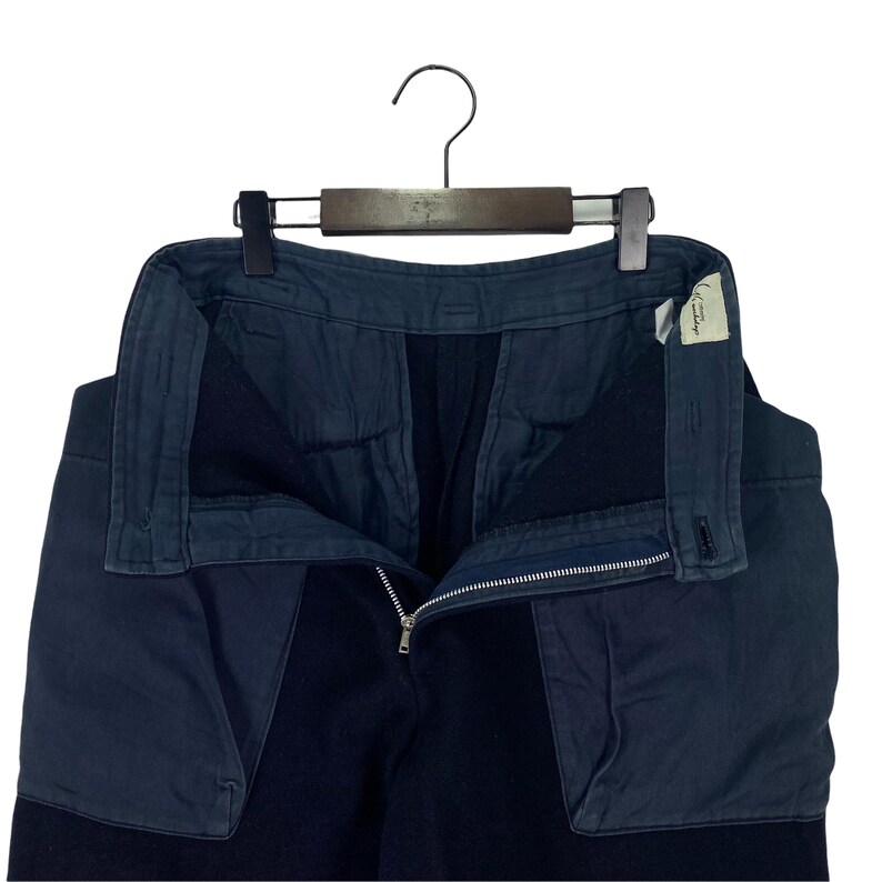 May include: Dark navy blue pants with a contrasting fabric panel on the front. The pants are hanging on a wooden hanger. The zipper is open, and a small tag is visible on the right side.