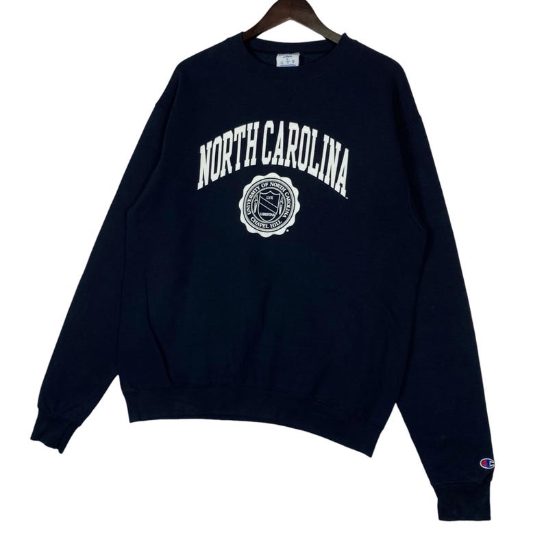 May include: Navy blue Champion sweatshirt with the words "NORTH CAROLINA" in white arched lettering. Below the text is a white circular emblem with text that reads "UNIVERSITY OF NORTH CAROLINA CHAPEL HILL".