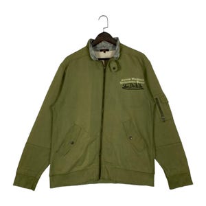 Puede incluir: Chaqueta verde oliva con cremallera, cuello gris y el texto "Custom Machine's Performance Parts Von Dutch". La chaqueta tiene dos bolsillos delanteros y un bolsillo con cremallera en la manga izquierda.
