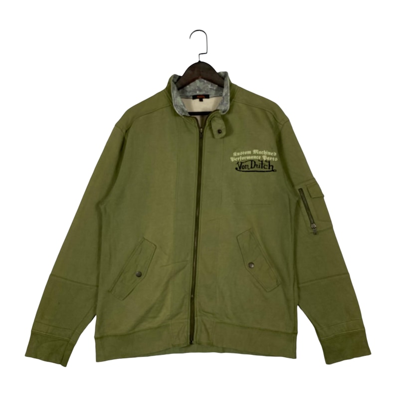 Puede incluir: Chaqueta verde oliva con cremallera, cuello gris y el texto "Custom Machine's Performance Parts Von Dutch". La chaqueta tiene dos bolsillos delanteros y un bolsillo con cremallera en la manga izquierda.