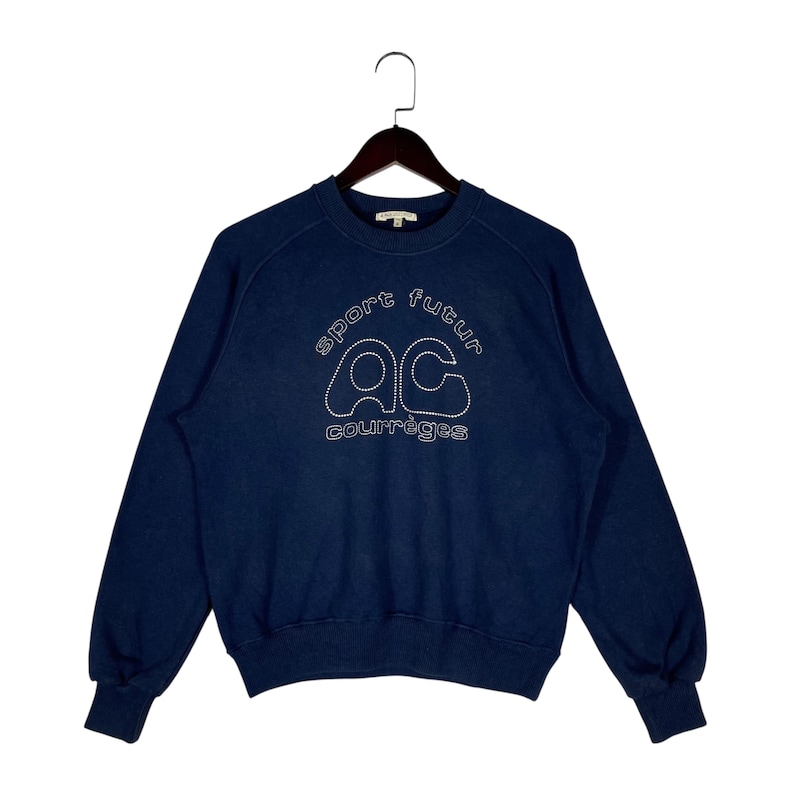 May include: Dark blue sweatshirt featuring white embroidered text and a logo. The text reads "sport futur" above the letters "AC" and the word "courr&egrave;ges" below. The sweatshirt is suspended from a wooden hanger.