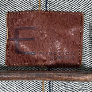 Puede incluir: Primer plano de una etiqueta de cuero marr&oacute;n con el texto en relieve "E FUNCTION EDWIN" sobre un fondo de mezclilla. La etiqueta es rectangular con esquinas redondeadas y un borde cosido.