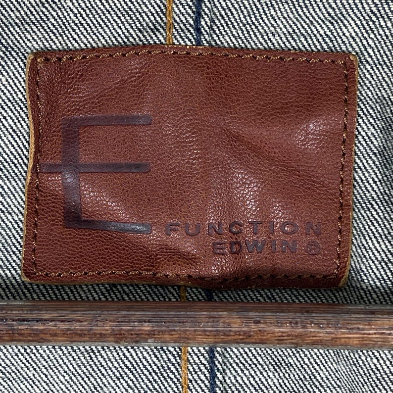 Puede incluir: Primer plano de una etiqueta de cuero marr&oacute;n con el texto en relieve "E FUNCTION EDWIN" sobre un fondo de mezclilla. La etiqueta es rectangular con esquinas redondeadas y un borde cosido.