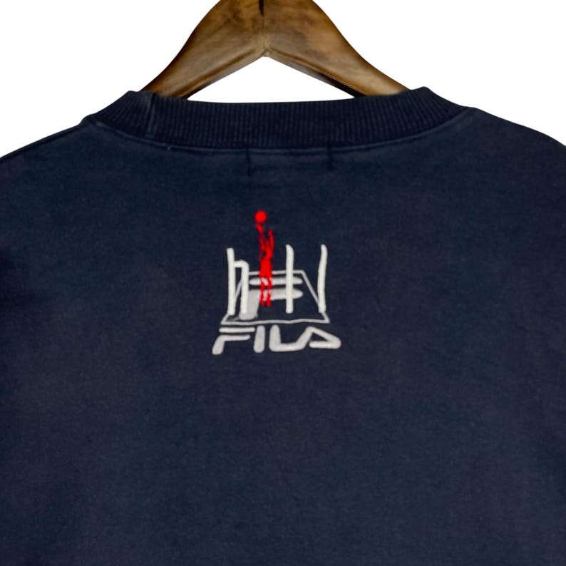 Puede incluir: Sudadera azul marino con un dise&ntilde;o bordado en blanco y rojo. El dise&ntilde;o incluye formas abstractas y el nombre de la marca "FILA" en blanco. La sudadera es de material suave y tiene cuello redondo.