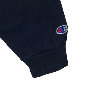 May include: Close-up of a navy blue sweatshirt sleeve. The cuff is ribbed and dark blue. A Champion logo is embroidered on the upper sleeve in red, white, and blue.