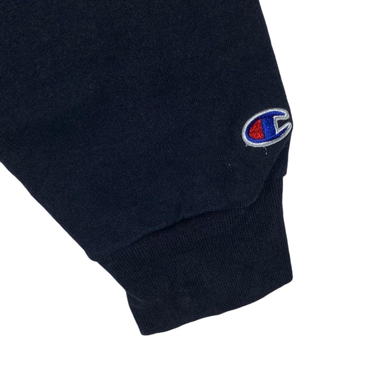 May include: Close-up of a navy blue sweatshirt sleeve. The cuff is ribbed and dark blue. A Champion logo is embroidered on the upper sleeve in red, white, and blue.