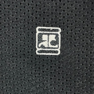 Pu&ograve; includere: Primo piano di un tessuto grigio scuro perforato. Una piccola toppa ricamata bianca &egrave; centrata sul tessuto. La toppa presenta un design stilizzato all'interno di un bordo rettangolare, che suggerisce un logo o un emblema.