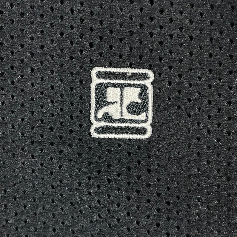 Pu&ograve; includere: Primo piano di un tessuto grigio scuro perforato. Una piccola toppa ricamata bianca &egrave; centrata sul tessuto. La toppa presenta un design stilizzato all'interno di un bordo rettangolare, che suggerisce un logo o un emblema.