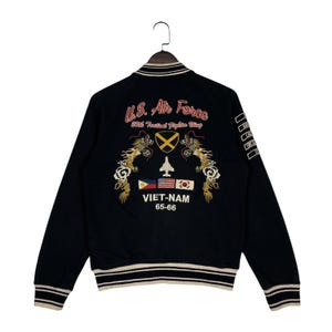 May include: Black bomber jacket with embroidered details. Features "U.S. Air Force" text, a fighter jet, and flags. The jacket has a striped collar, cuffs, and hem. The text "VIET-NAM 65-66" is also visible.