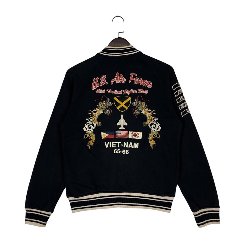 May include: Black bomber jacket with embroidered details. Features "U.S. Air Force" text, a fighter jet, and flags. The jacket has a striped collar, cuffs, and hem. The text "VIET-NAM 65-66" is also visible.