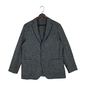 Puede incluir: Un blazer de tweed gris con un patrón de cuadros azules, colgado de una percha de madera. La chaqueta tiene dos bolsillos delanteros y un cierre de dos botones. El blazer es de estilo clásico, adecuado para diversas ocasiones.
