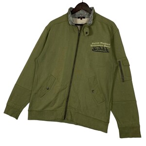 Puede incluir: Chaqueta verde oliva con cremallera, cuello alzado y forro gris en contraste. Cuenta con dos bolsillos delanteros, un bolsillo con cremallera en la manga y el texto "Custom Machines Performance Parts Von Dutch" bordado en el pecho.