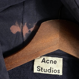 May include: Close-up of a navy blue hoodie with a brown stain on the hood. The hoodie is hanging on a wooden hanger. A white tag with the text "Acne Studios" is visible.
