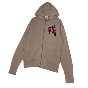 Puede incluir: Una sudadera con capucha y cremallera de color beige con capucha y bolsillos delanteros. La sudadera con capucha presenta un gr&aacute;fico en el pecho con el texto "E.V.I.S.U. TRACK" en rojo y azul marino. Las mangas tienen pu&ntilde;os acanalados.