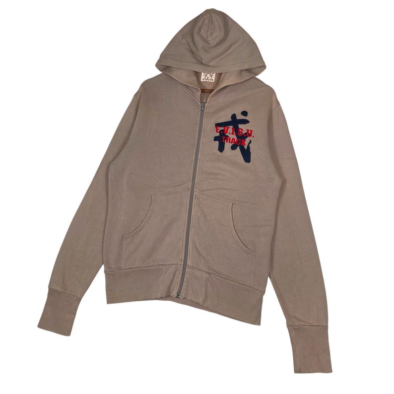 Puede incluir: Una sudadera con capucha y cremallera de color beige con capucha y bolsillos delanteros. La sudadera con capucha presenta un gr&aacute;fico en el pecho con el texto "E.V.I.S.U. TRACK" en rojo y azul marino. Las mangas tienen pu&ntilde;os acanalados.