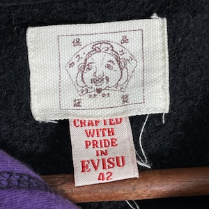 May include: Close-up of a white tag with a detailed illustration and text. The tag reads "CRAFTED WITH PRIDE IN EVISU 42" in red. The background is black, with a glimpse of purple fabric.