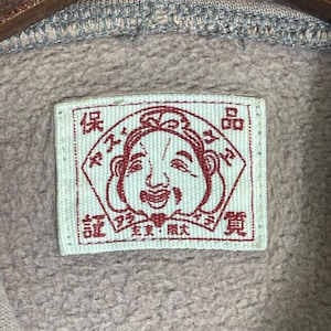 Puede incluir: Primer plano de una etiqueta rectangular con una ilustraci&oacute;n de una cara sonriente en tinta roja. La etiqueta est&aacute; sobre una tela marr&oacute;n claro. Caracteres japoneses rodean la cara.