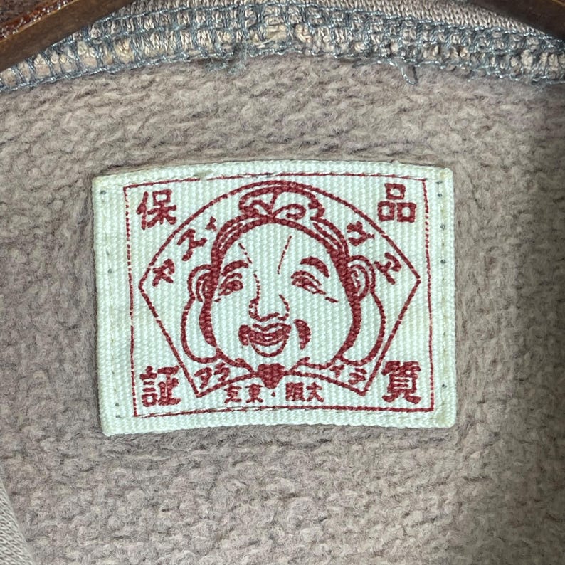 Puede incluir: Primer plano de una etiqueta rectangular con una ilustraci&oacute;n de una cara sonriente en tinta roja. La etiqueta est&aacute; sobre una tela marr&oacute;n claro. Caracteres japoneses rodean la cara.