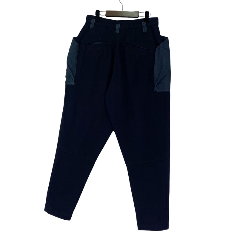 May include: Dark navy blue pants with two large pockets on the sides. The pants are hanging on a wooden hanger. The pockets are a contrasting dark blue color. The pants have a high waist and a relaxed fit.