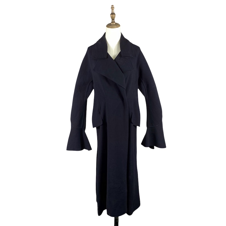 May include: A long, dark navy coat with a unique design. The coat features a wrap-style front, wide lapels, and flared sleeves. It is displayed on a mannequin against a white background, showcasing its full length and tailored silhouette.