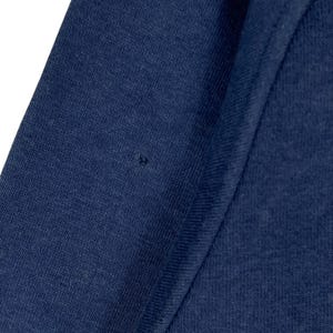May include: A close-up shot of navy blue fabric, likely from an item of clothing. The material has a textured surface, with a small dark mark present. The image focuses on the colour and weave of the fabric.