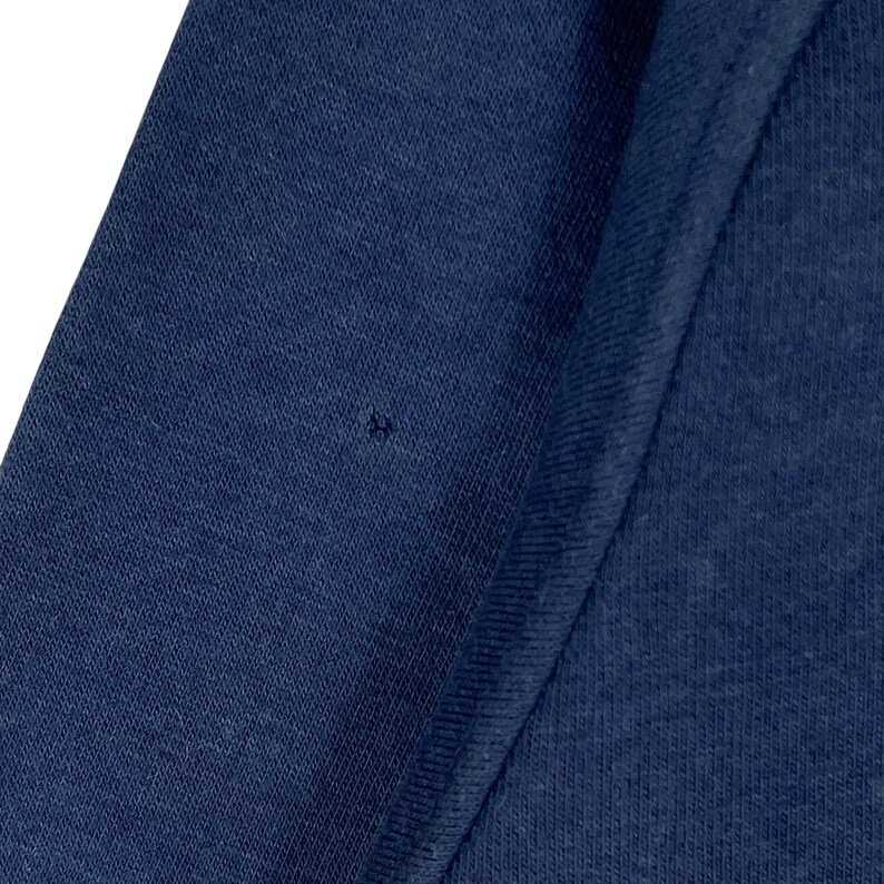 May include: A close-up shot of navy blue fabric, likely from an item of clothing. The material has a textured surface, with a small dark mark present. The image focuses on the colour and weave of the fabric.