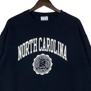 May include: Navy blue sweatshirt with the words "NORTH CAROLINA" in white arched lettering. Below the text is a circular emblem with the words "UNIVERSITY OF NORTH CAROLINA CHAPEL HILL" and the word "LIBERTAS".