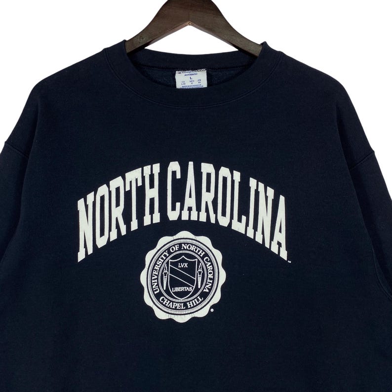 May include: Navy blue sweatshirt with the words "NORTH CAROLINA" in white arched lettering. Below the text is a circular emblem with the words "UNIVERSITY OF NORTH CAROLINA CHAPEL HILL" and the word "LIBERTAS".