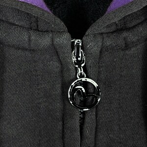 May include: Close-up of a black hooded sweatshirt with a silver zipper and a circular zipper pull. The zipper pull has a black and white logo. The inside of the hood is purple.