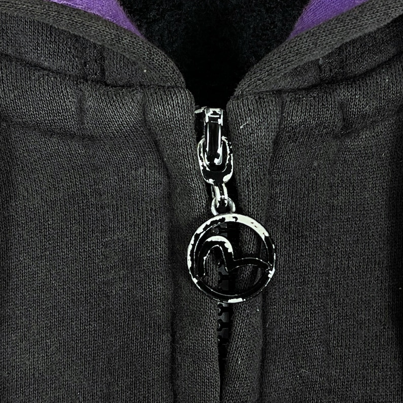 May include: Close-up of a black hooded sweatshirt with a silver zipper and a circular zipper pull. The zipper pull has a black and white logo. The inside of the hood is purple.