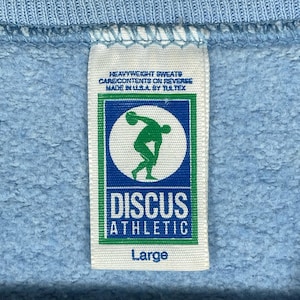 May include: A white fabric clothing tag sewn onto a light blue sweatshirt. The tag features the text HEAVYWEIGHT SWEATS, CARE CONTENTS ON REVERSE, MADE IN U.S.A. BY TULTEX, DISCUS ATHLETIC, and Large, with a green and blue logo of a discus thrower.