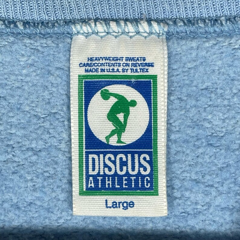 May include: A white fabric clothing tag sewn onto a light blue sweatshirt. The tag features the text HEAVYWEIGHT SWEATS, CARE CONTENTS ON REVERSE, MADE IN U.S.A. BY TULTEX, DISCUS ATHLETIC, and Large, with a green and blue logo of a discus thrower.
