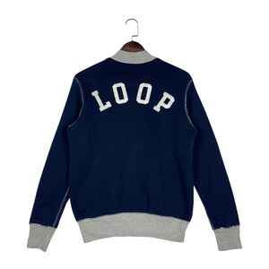 May include: A dark navy blue long-sleeve sweatshirt featuring a light gray ribbed collar, cuffs, and hem. The back of the garment displays the word LOOP in large, white, block-style capital letters centered across the upper back area.