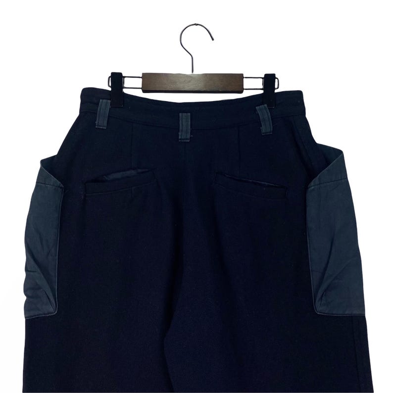 May include: Dark navy blue pants with two large pockets on the back, and two smaller pockets. The pants are hanging on a black hanger against a white background. The pockets are a darker shade of blue.