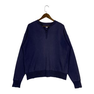 May include: A dark navy blue long-sleeve sweatshirt featuring a V-neck collar detail and a ribbed hemline. The garment has long sleeves with ribbed cuffs and is displayed on a wooden hanger, showcasing a solid color design throughout the piece.
