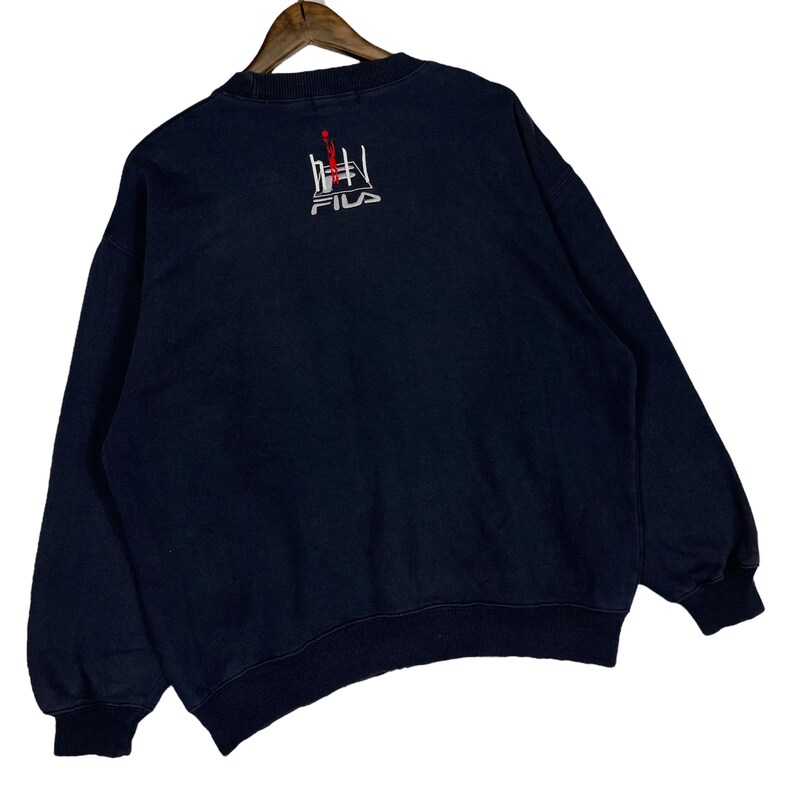 Puede incluir: Sudadera azul marino con cuello redondo y mangas largas. La parte trasera presenta un dise&ntilde;o bordado en rojo y blanco sobre el logotipo de FILA. La sudadera est&aacute; colgada en una percha de madera.