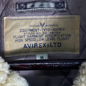 May include: A close-up of a jacket label with the text "EQUIPMENT-TYPE-AVIREX" and "AVIREX LTD". The label is beige with black text and a blue emblem. The jacket has a quilted lining and a cream-coloured, fluffy collar.