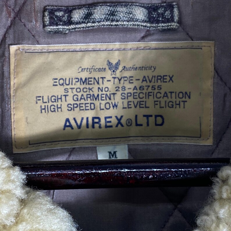 May include: A close-up of a jacket label with the text "EQUIPMENT-TYPE-AVIREX" and "AVIREX LTD". The label is beige with black text and a blue emblem. The jacket has a quilted lining and a cream-coloured, fluffy collar.